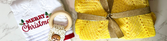 A holiday-themed crafting scene featuring a white towel with 'Merry Christmas' embroidered on it and a yellow crocheted toddler dress tied with a ribbon, all resting on a marble surface.